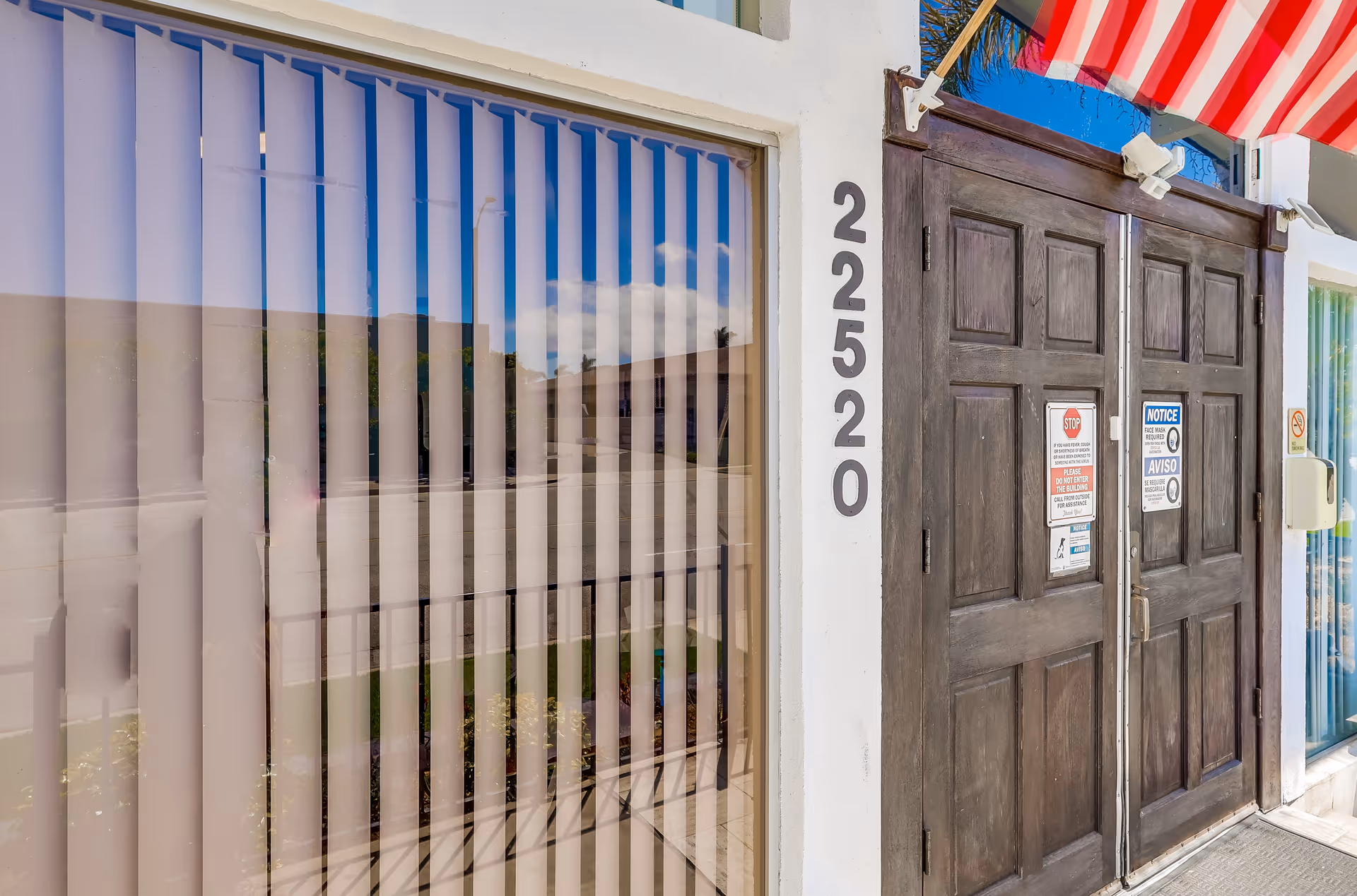 Entrance to a building with dark wooden double doors and vertical blinds covering a large window next to the door. The building number 22520 is displayed vertically on the wall beside the door. An American flag is partially visible above the entrance.