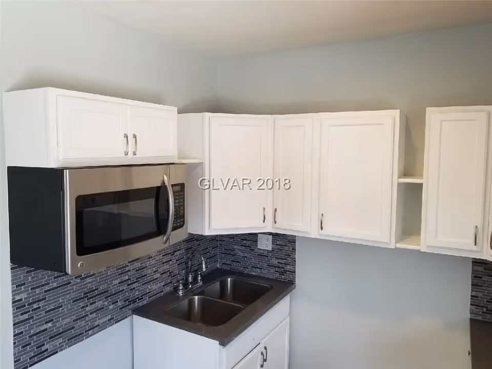 A modern kitchen corner featuring white upper cabinets, a stainless steel microwave mounted above a black countertop with a double sink, and a gray and black tiled backsplash.