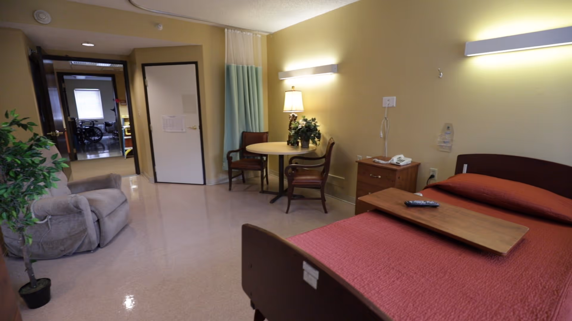 A nursing facility room with a single bed covered in a red blanket, a wooden bedside table with a telephone, a small round table with two chairs, a lamp, and a potted plant. There is an armchair on the left side and a doorway leading to another room with a wheelchair visible.