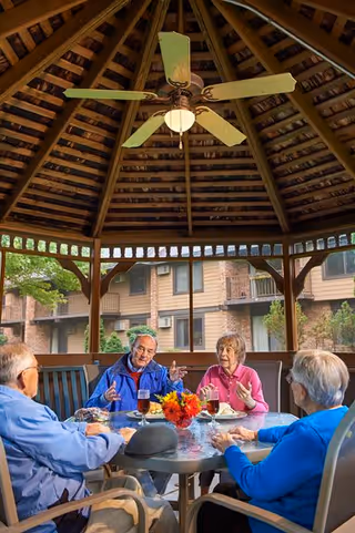 Four elderly people sitting around a round glass table under a wooden gazebo with a ceiling fan, engaged in conversation and enjoying drinks. The background shows a residential building with balconies and greenery.