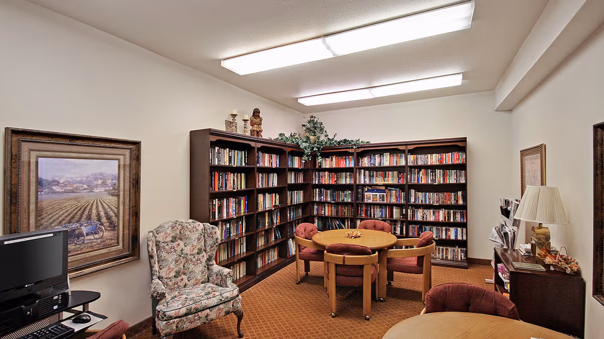 A cozy library room with corner bookshelves filled with books, a round wooden table surrounded by four cushioned chairs, a floral upholstered armchair, a computer desk with a monitor and keyboard, framed artwork on the walls, and a lamp on a side table.
