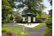 Park-like outdoor courtyard with paved paths, informational kiosks and small gazebo structures surrounded by trees and grass.