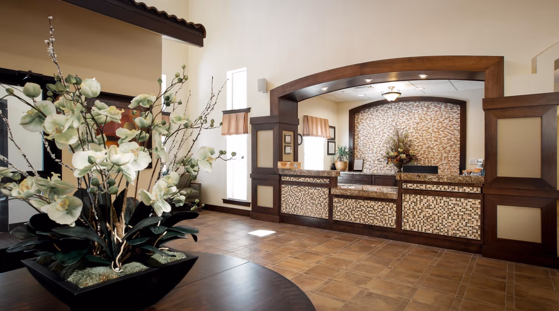 Reception area of a senior living facility with a mosaic-tiled front desk, wooden trim, and a large floral arrangement on a round table in the foreground. The space is well-lit with natural light coming through tall windows with beige curtains.