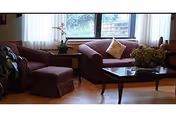 Sunlit sitting area with two upholstered sofas, an ottoman, a coffee table with flowers, and a large window with curtains.