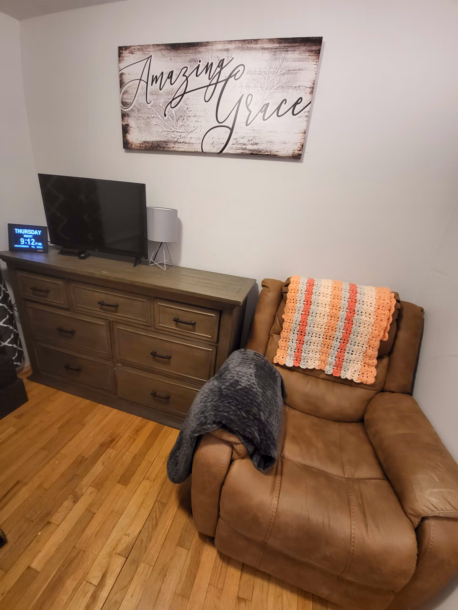 A cozy corner of a room featuring a brown leather recliner chair with a crocheted blanket draped over the back and a dark gray throw on one arm. Next to the chair is a wooden dresser with multiple drawers, on top of which sits a flat-screen TV, a small lamp, and a digital clock displaying the day and time. Above the dresser hangs a decorative wall sign that reads 'Amazing Grace'. The room has hardwood flooring and light-colored walls.