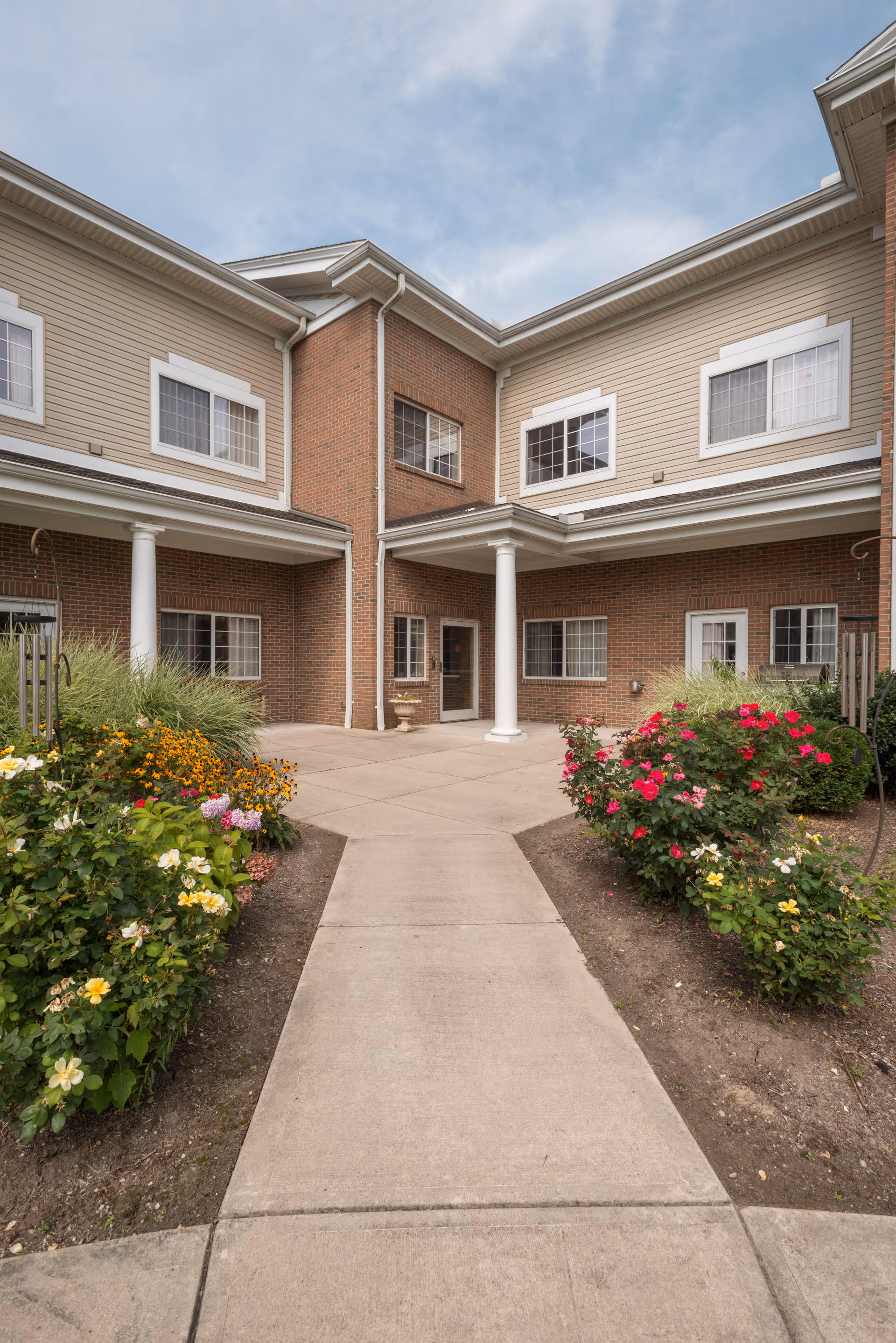 Concrete walkway leading to the entrance of a two-story brick-and-siding building with columns, windows, and flower beds.