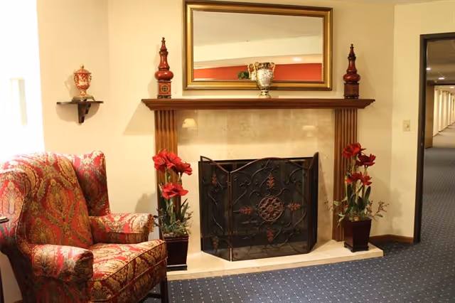 A cozy interior space featuring a decorative fireplace with a black wrought iron screen, flanked by two potted red flower plants. Above the fireplace is a large rectangular mirror with a wooden frame. To the left of the fireplace is a patterned upholstered armchair in red and gold tones. The walls are light-colored, and the floor is carpeted in a dark blue with small white dots. There is a hallway visible to the right.