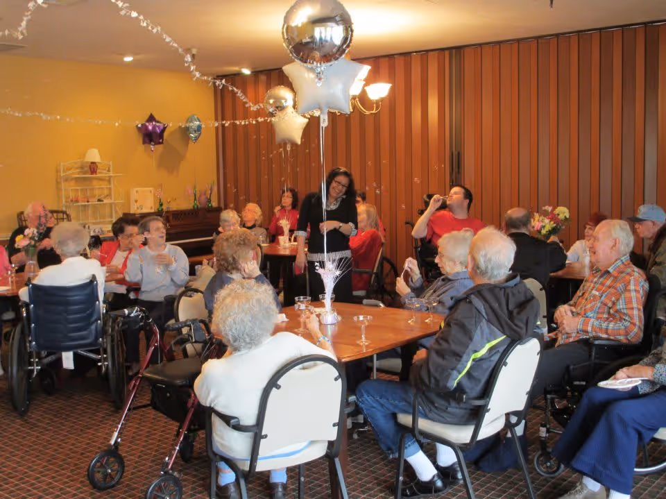 Seniors seated around tables in a decorated common room with balloons celebrating together.