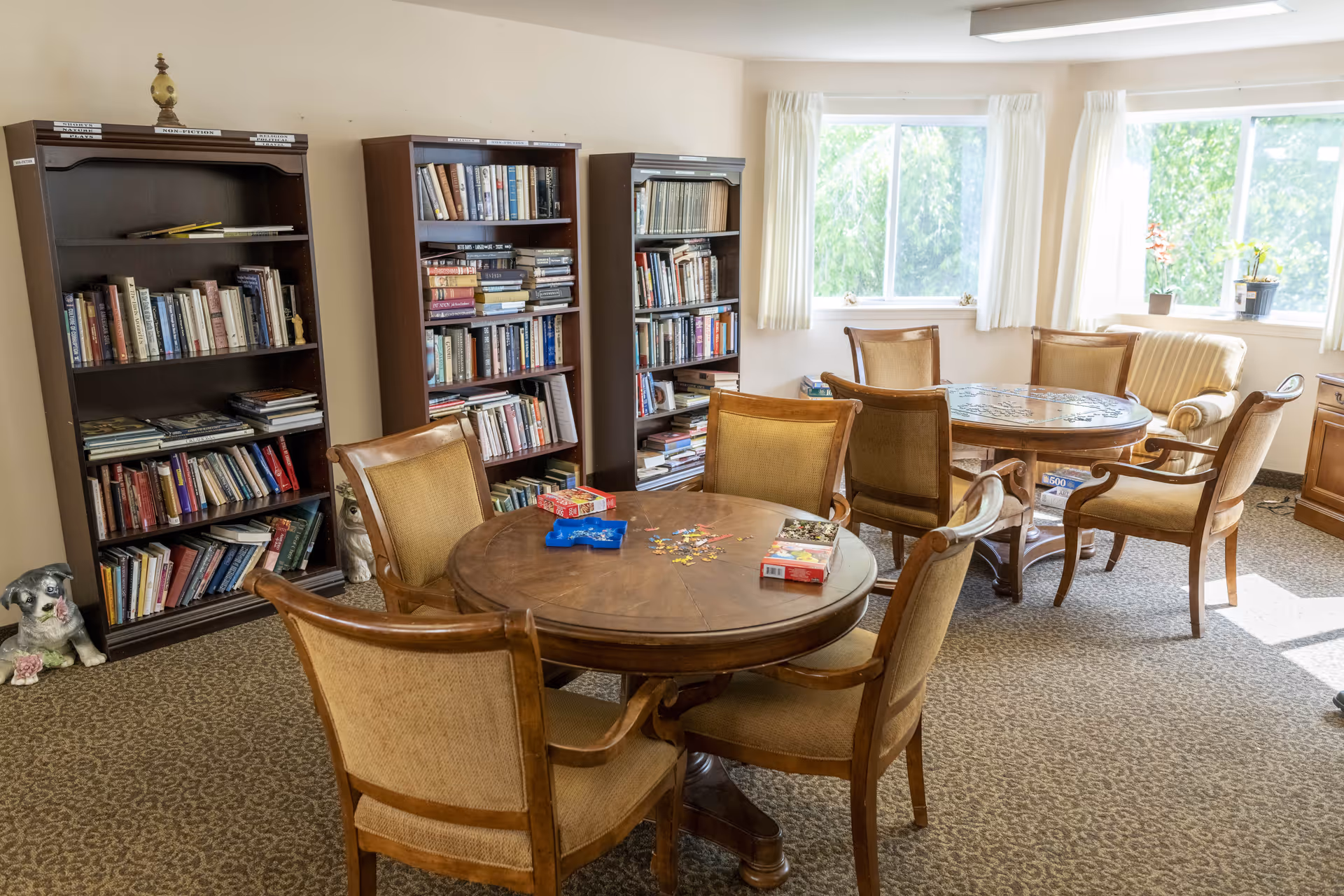 A cozy room with two round wooden tables surrounded by cushioned wooden chairs. On one table, there is a partially completed jigsaw puzzle and some puzzle boxes. Behind the tables, there are three tall bookshelves filled with books. The room has large windows with white curtains letting in natural light, and a beige carpet covers the floor. There are also small decorative items including a dog figurine on the floor near the bookshelves.