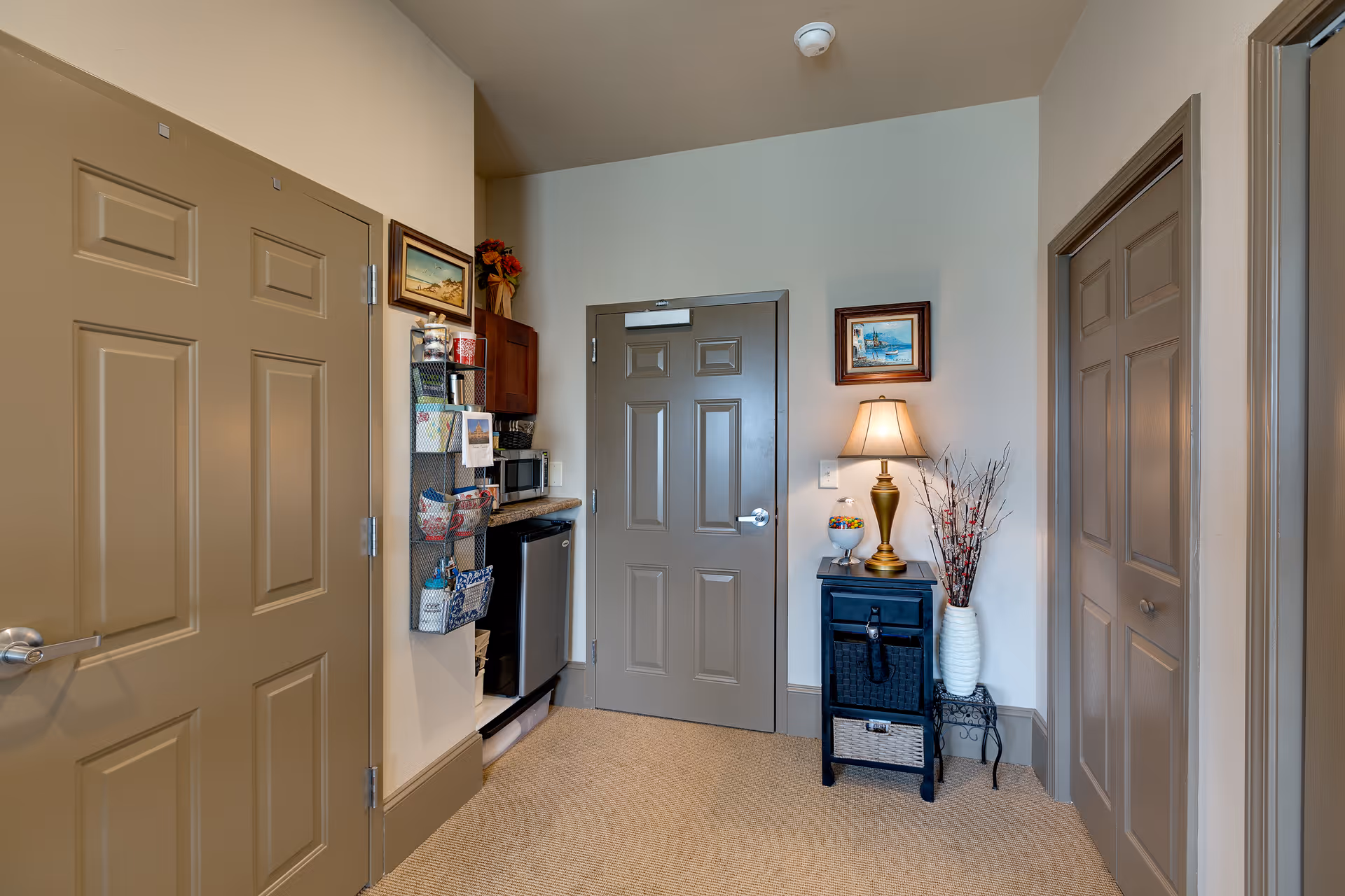 Interior view of a senior living facility room corner with three closed beige doors, a small black cabinet with a lamp and decorative vase on top, a wall-mounted organizer with various items, a mini refrigerator, and a microwave on a countertop.