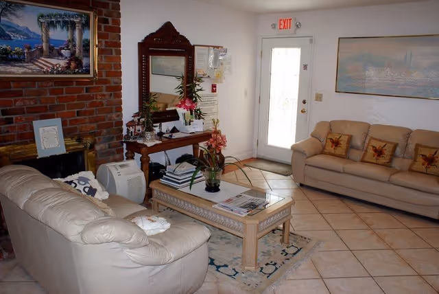 A cozy living room with two beige leather sofas, a wooden coffee table with a glass top, and a decorative rug. There is a brick fireplace on the left side with a framed painting above it. A wooden console table with a large mirror and various decorative items is against the wall near a white door with an exit sign above it. The floor is tiled, and there is a large framed artwork on the wall above one of the sofas.