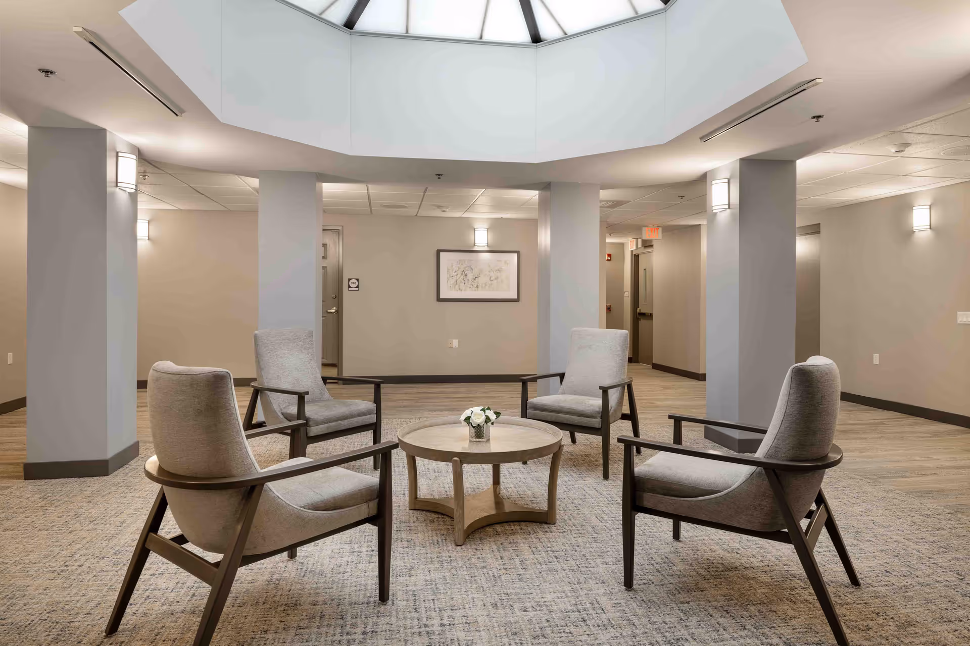 A modern senior living community common area with four gray upholstered armchairs arranged around a round wooden coffee table with a small flower arrangement. The room features neutral-colored walls, carpeted flooring, several columns, and a large skylight overhead providing natural light.