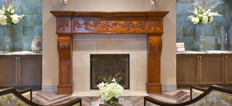 A cozy living room area featuring a decorative wooden fireplace mantel with intricate tree carvings. On either side of the fireplace are built-in cabinets with granite countertops, each adorned with a tall vase of white flowers and decorative items. The floor has a patterned wood design.