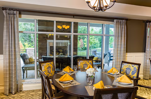 Round dining table set with yellow napkins in a dining room facing large windows and an outdoor seating area.