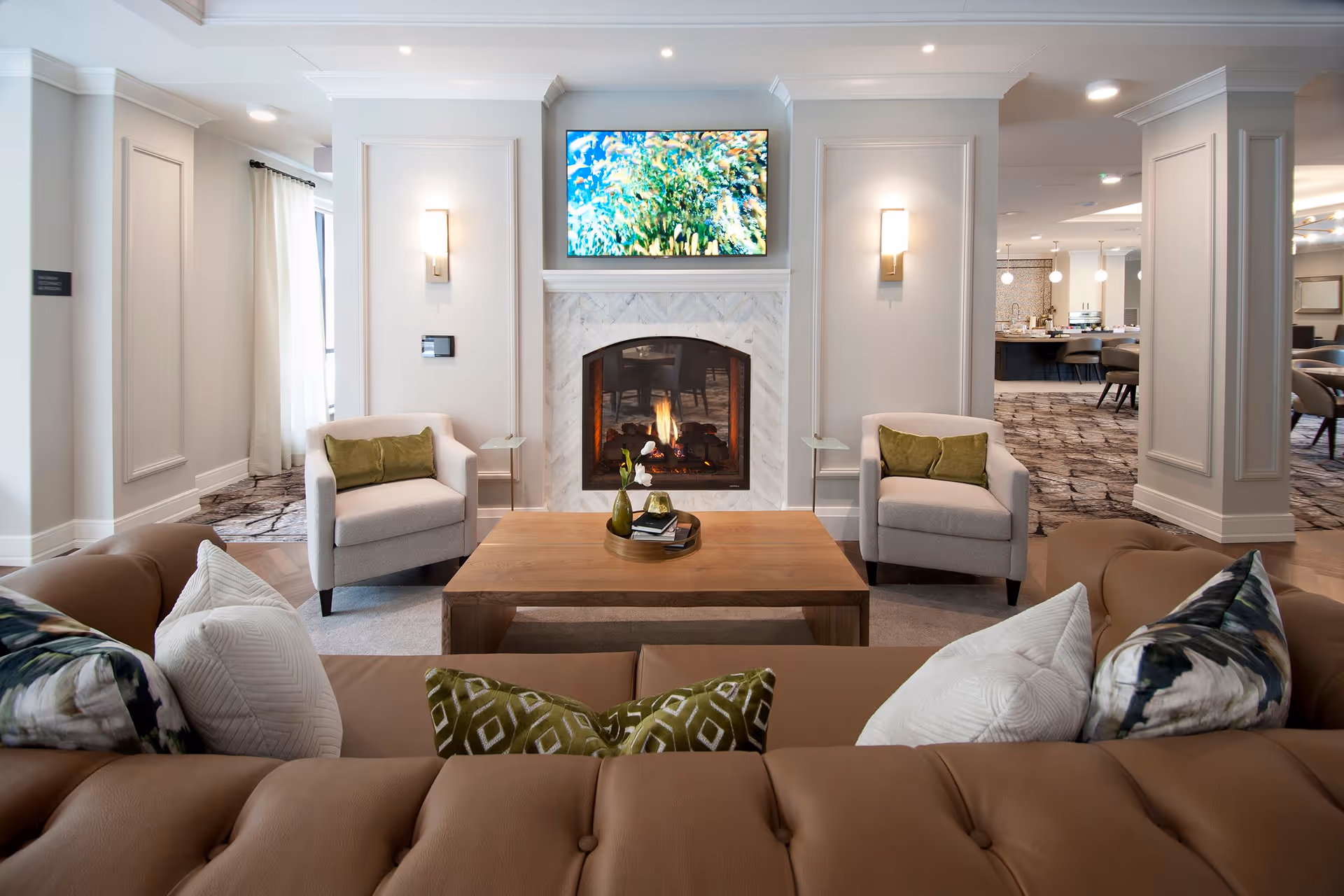 A cozy living room area with a brown leather sofa adorned with patterned pillows, two beige armchairs with green cushions, and a wooden coffee table with decorative items. A fireplace with a lit fire is centered on the far wall, above which a flat-screen TV displays a colorful image. The room has light-colored walls with panel molding and wall sconces, and an open view into a dining area with tables and chairs.