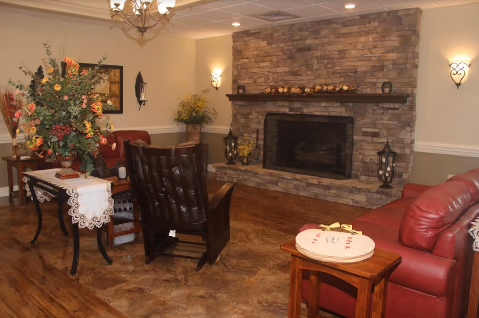 Cozy common room with a stone fireplace, leather seating, wooden tables and a large floral arrangement.