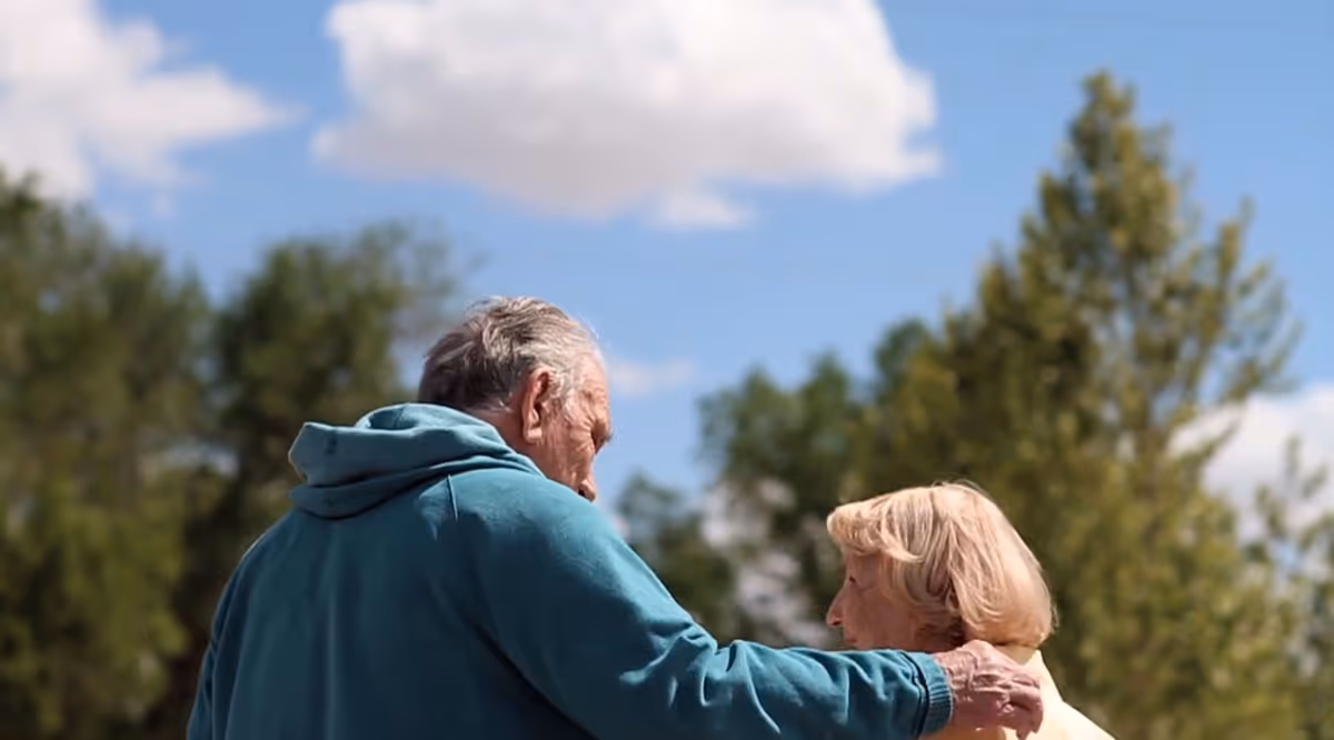 An elderly man and woman outdoors with the man’s arm around the woman’s shoulder, trees and a partly cloudy sky in the background.