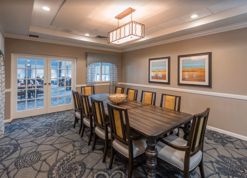 A dining room with a long wooden table surrounded by ten chairs with cushioned seats and backs. The room has beige walls with white trim, two framed landscape paintings on the wall, a window with patterned curtains, and a modern rectangular ceiling light fixture. French doors with glass panes lead to another room with additional seating.