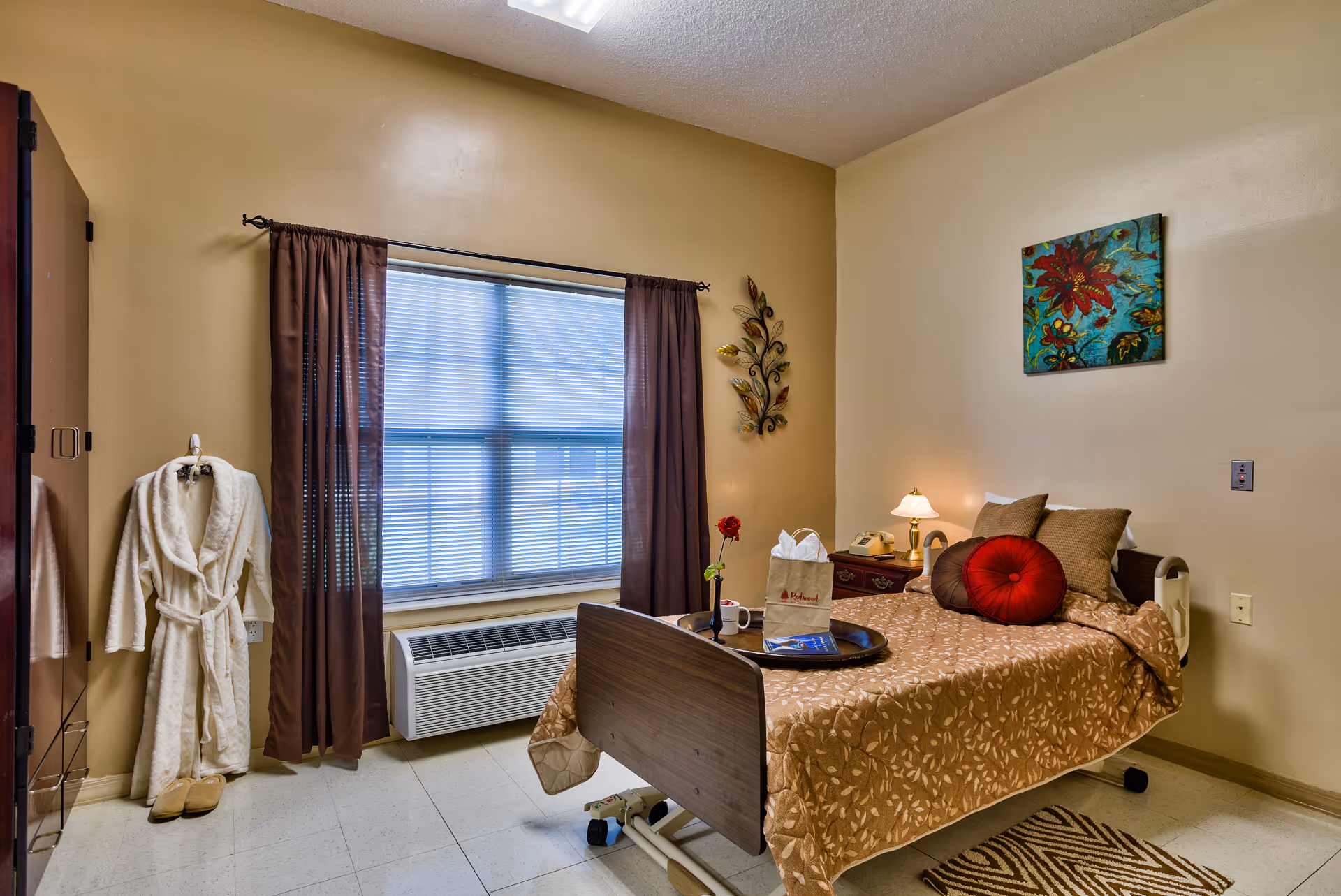A cozy bedroom in a senior living facility featuring a single bed with a brown patterned bedspread, decorative pillows, and a tray with a rose and a paper bag on it. There is a nightstand with a lamp and a telephone beside the bed. A window with brown curtains and blinds is on the wall, and a white bathrobe with slippers hangs on a hook near the door. The walls are beige with a colorful floral painting and a metal leaf wall decoration.