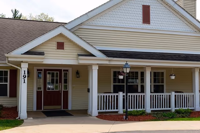 Front exterior view of a beige building with white trim and a covered porch featuring white railings and hanging flower pots. The entrance has double doors painted in dark red with glass panels, and the building number 191 is displayed on a white column near the door.