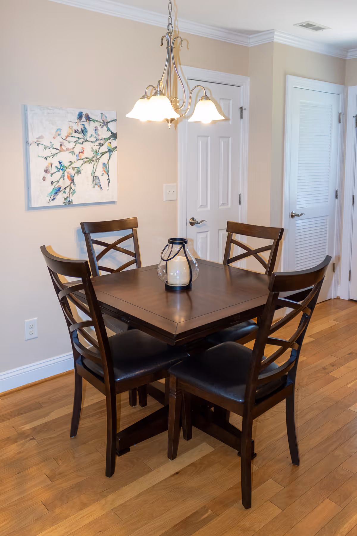 A small dining area with a square wooden table and four matching wooden chairs with black cushioned seats. A decorative lantern with a candle is placed in the center of the table. Above the table hangs a chandelier with three lights. On the wall behind the table is a painting of colorful birds on branches. The room has light-colored walls, wooden flooring, and two white doors in the background.