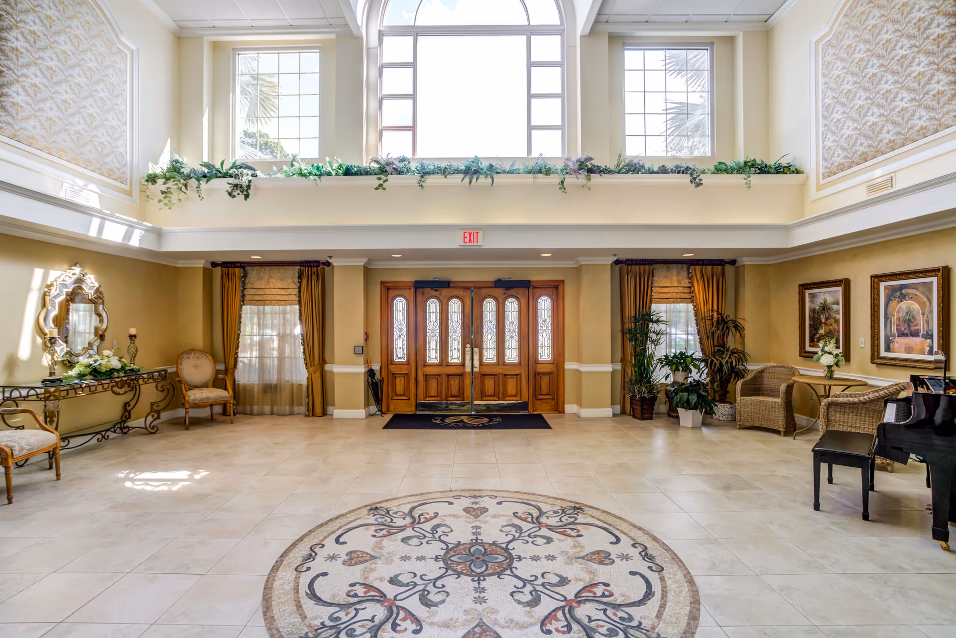 Spacious and elegant lobby area with large wooden double doors, tall windows letting in natural light, decorative plants on a ledge above the doors, a patterned circular rug on the tiled floor, ornate mirror and chairs on the left, and wicker chairs with framed artwork on the right.