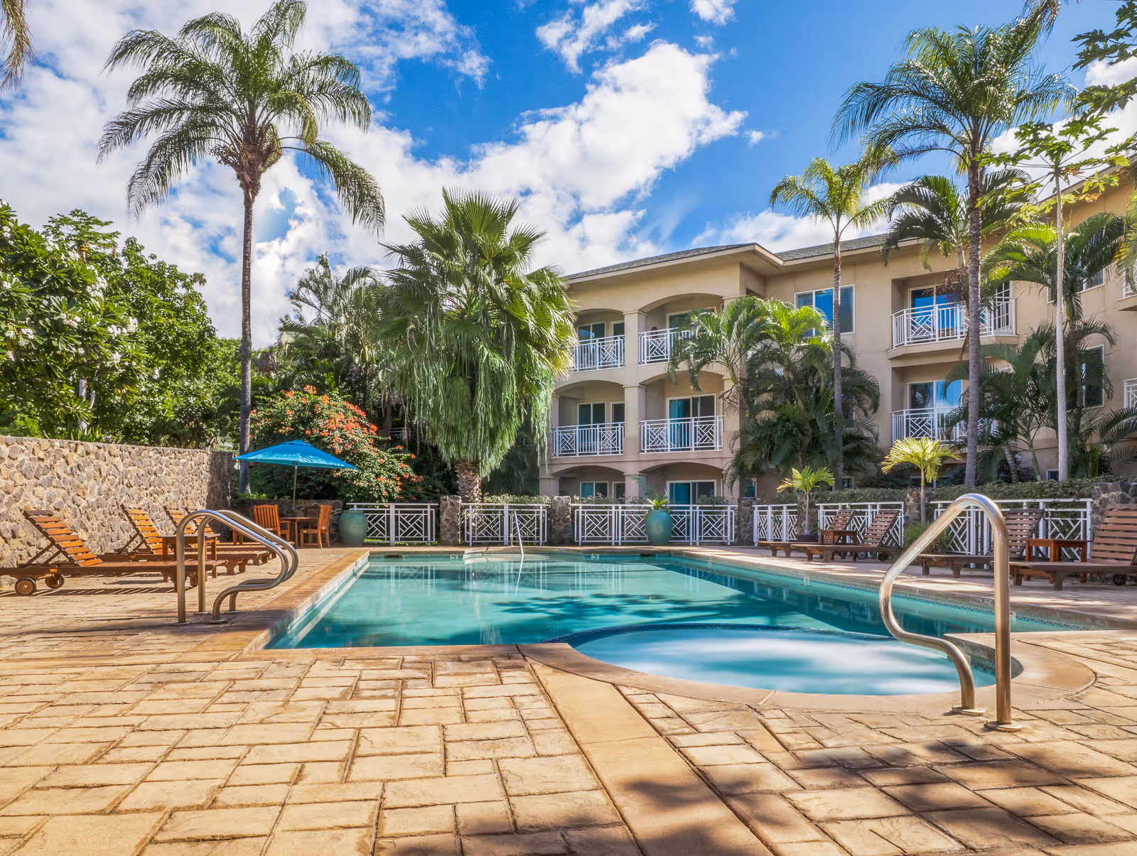 Outdoor swimming pool area at Holiday Kalama Heights with lounge chairs, palm trees, and a three-story building with balconies in the background under a partly cloudy blue sky.