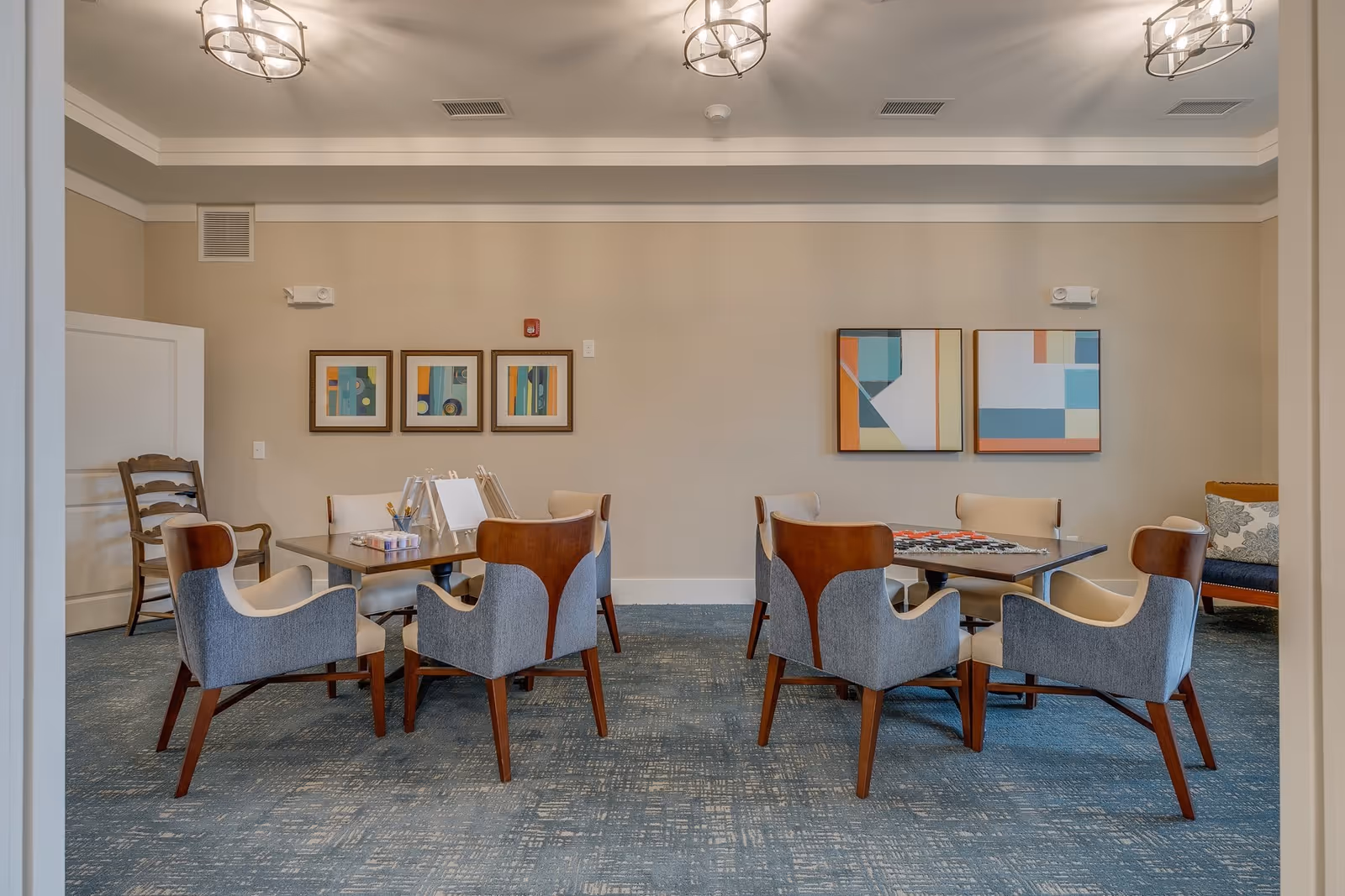 A bright common room with two square tables surrounded by upholstered chairs, abstract wall art, and overhead light fixtures.