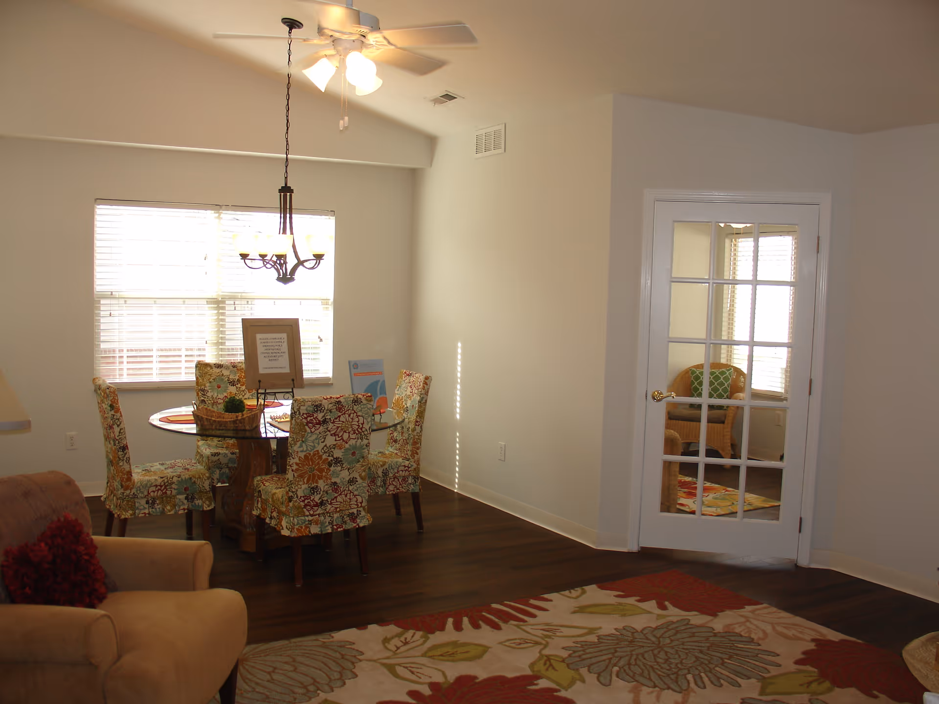 Interior view of a dining area with a round glass table surrounded by six floral upholstered chairs. A chandelier hangs above the table, and a window with blinds is behind it. To the right, there is a white door with glass panes leading to another room with a wicker chair and a green cushion. The floor is dark wood, and a floral patterned rug is partially visible in the foreground.
