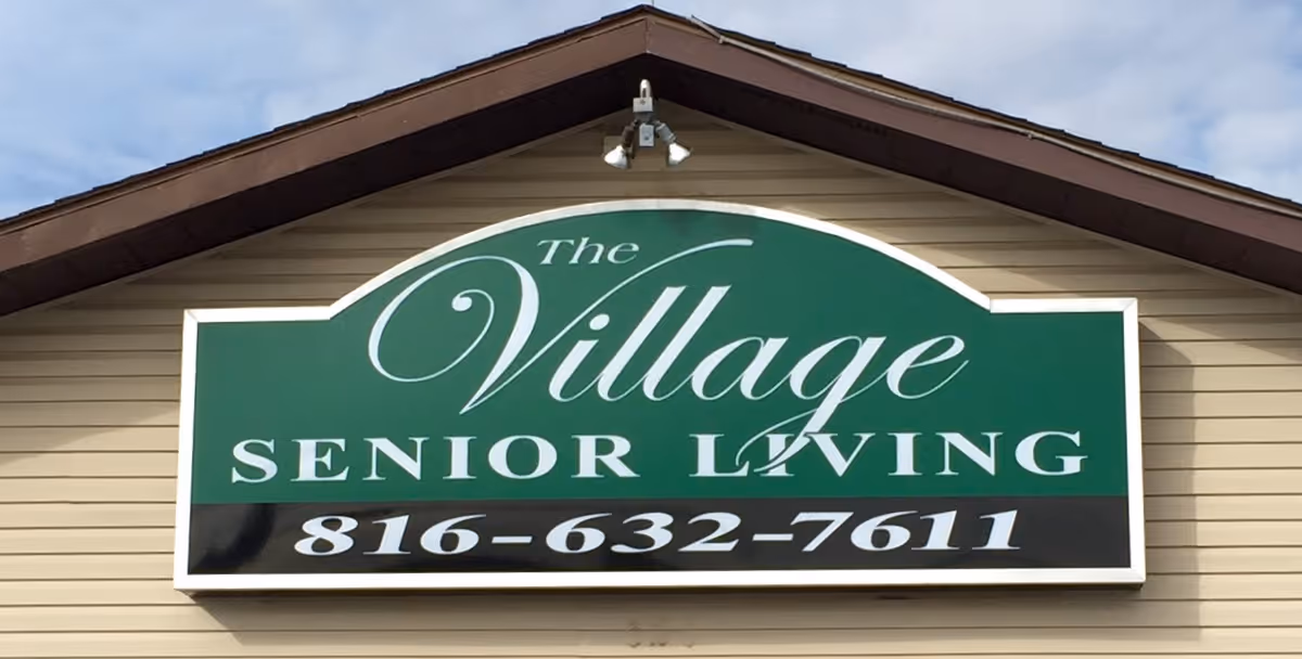 A green and white sign mounted on the exterior of a beige building with brown trim. The sign reads 'The Village Senior Living' with a phone number 816-632-7611 below.