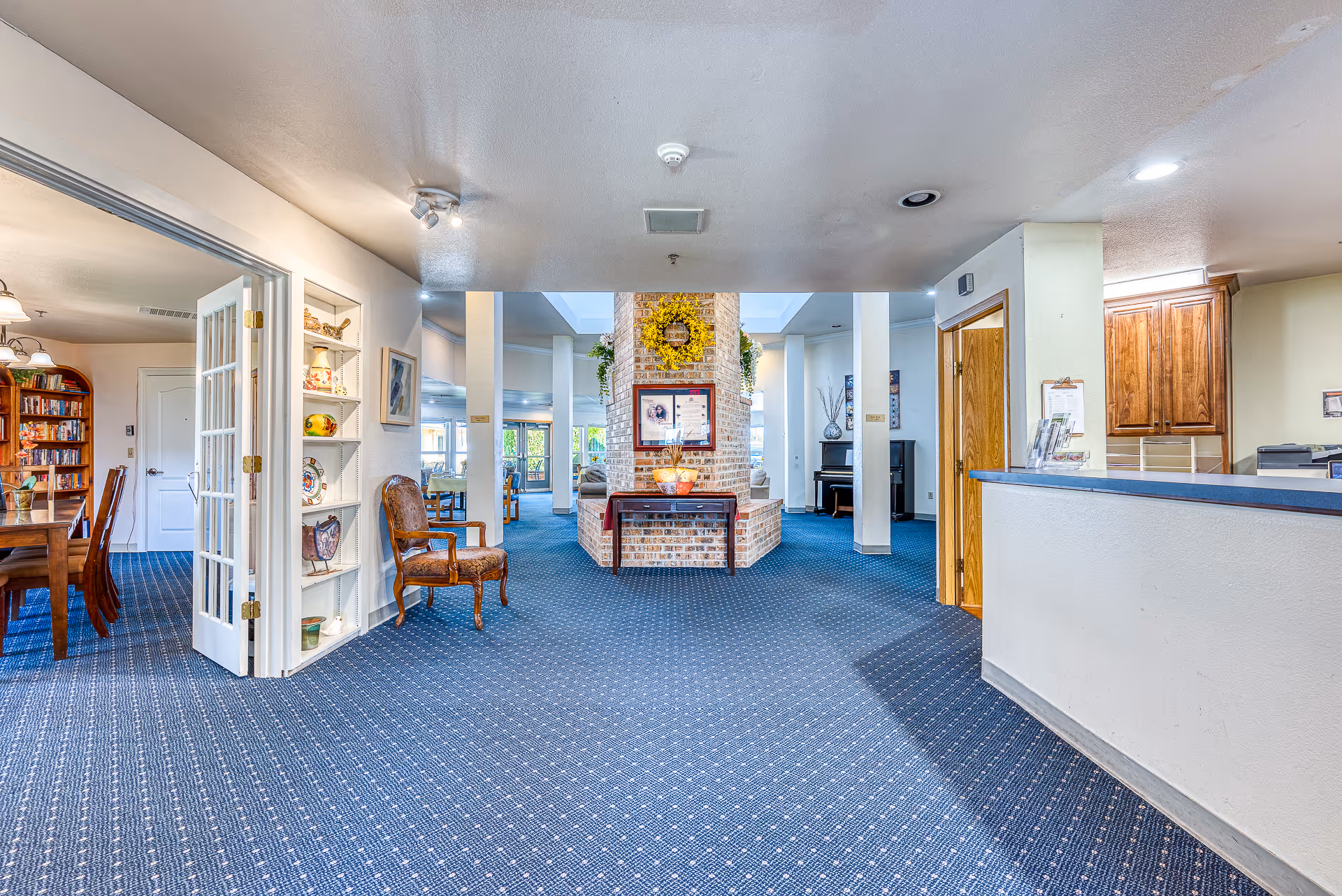 Spacious carpeted assisted living common area with a brick fireplace centerpiece, seating, a reception desk on the right and a dining room visible to the left.
