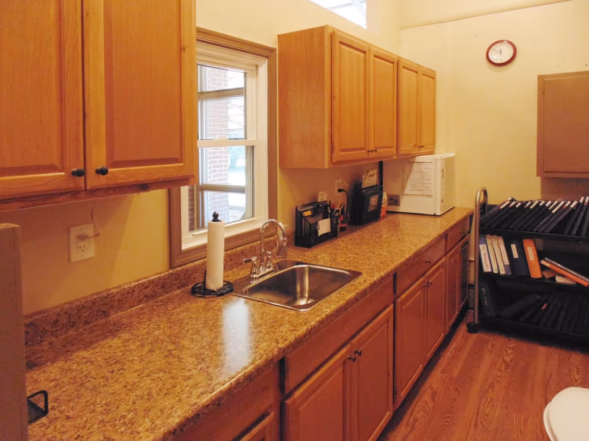 Interior view of a kitchen area with wooden cabinets, a countertop with a sink, a paper towel holder, a small microwave, and a rack filled with binders and folders. A window is above the sink, and a clock is mounted on the wall.