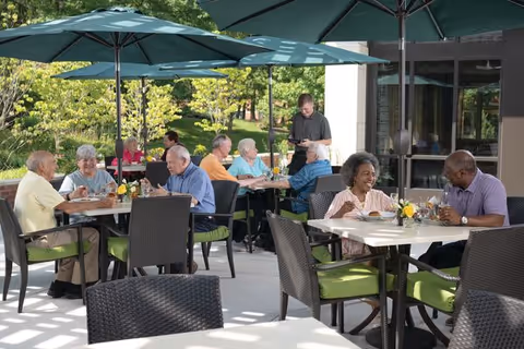 Seniors sitting at outdoor tables under large umbrellas, enjoying food and conversation at a senior living community patio. A server is taking an order from one of the tables. The setting is bright and surrounded by greenery.