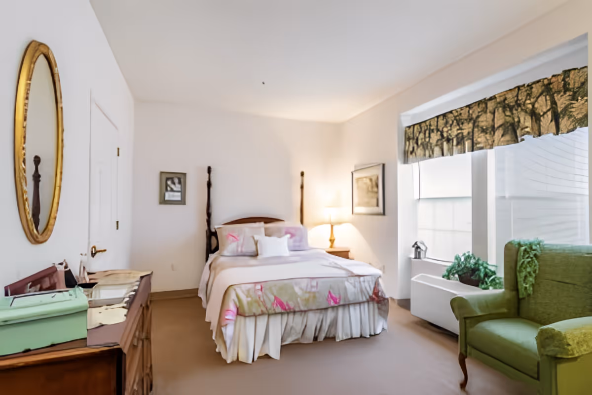 A cozy bedroom with a bed featuring a floral bedspread and multiple pillows, a wooden dresser with a green box on top, a round gold-framed mirror on the wall, a green upholstered armchair near a window with a patterned valance, and a bedside table with a lamp and framed artwork on the walls.