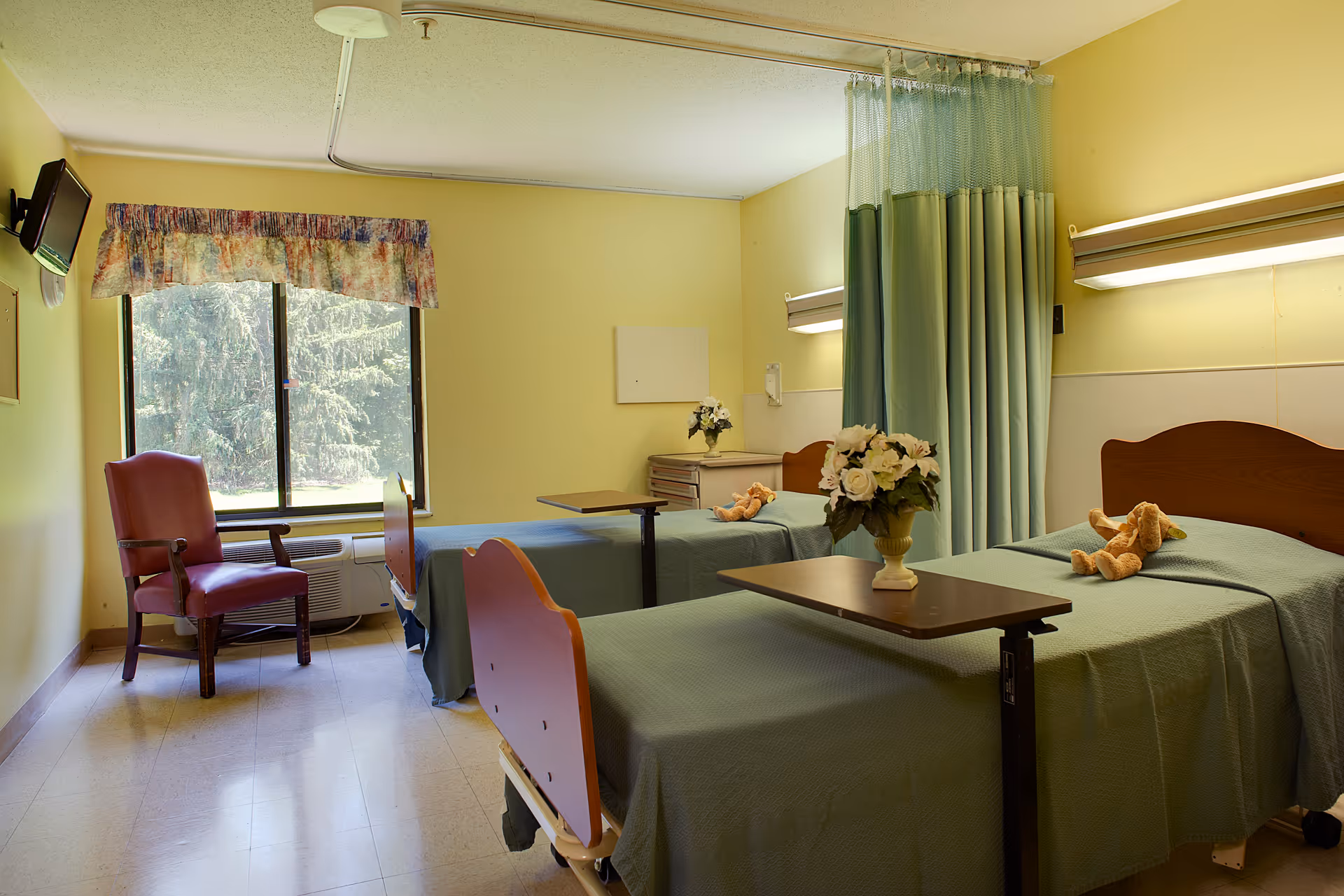 A room with two hospital-style beds separated by a green privacy curtain. Each bed has a green bedspread and a small teddy bear on it. There is a small table with a flower vase on the bed in the foreground. A red armchair is placed near a window with a floral valance, and a wall-mounted TV is visible on the left side. The walls are painted yellow, and the floor is tiled.