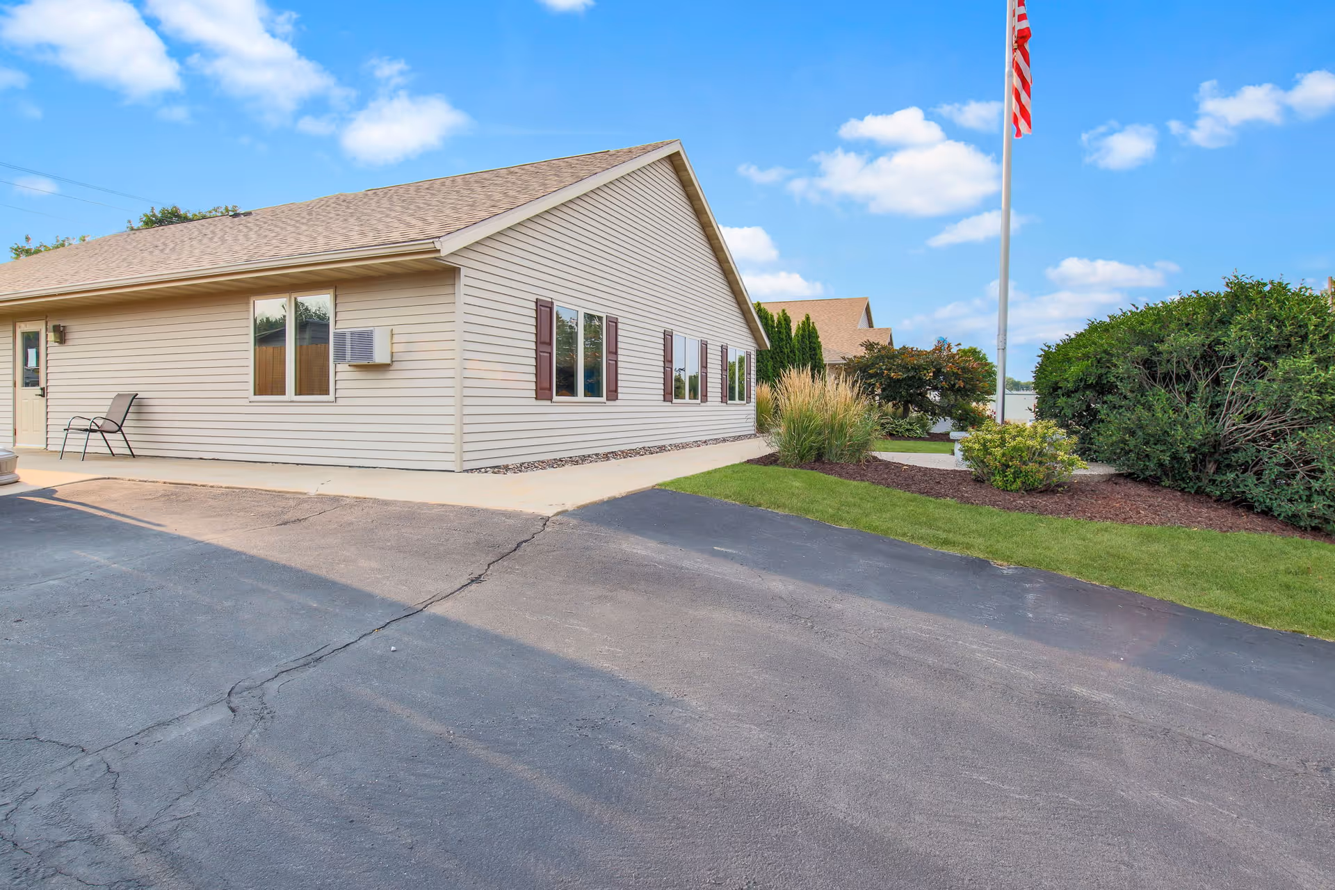 Exterior view of a single-story beige building with brown shutters, a small air conditioning unit, and a chair near the entrance. The building is surrounded by a paved driveway, green grass, bushes, and a flagpole with an American flag under a partly cloudy blue sky.