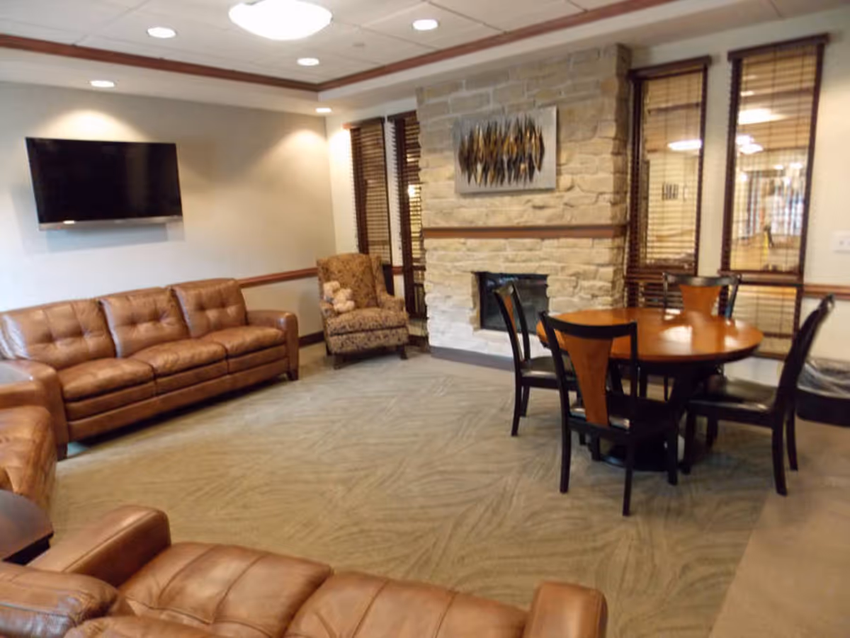 Communal lounge with leather sofas, a wall-mounted TV, a stone fireplace, and a round table with chairs.