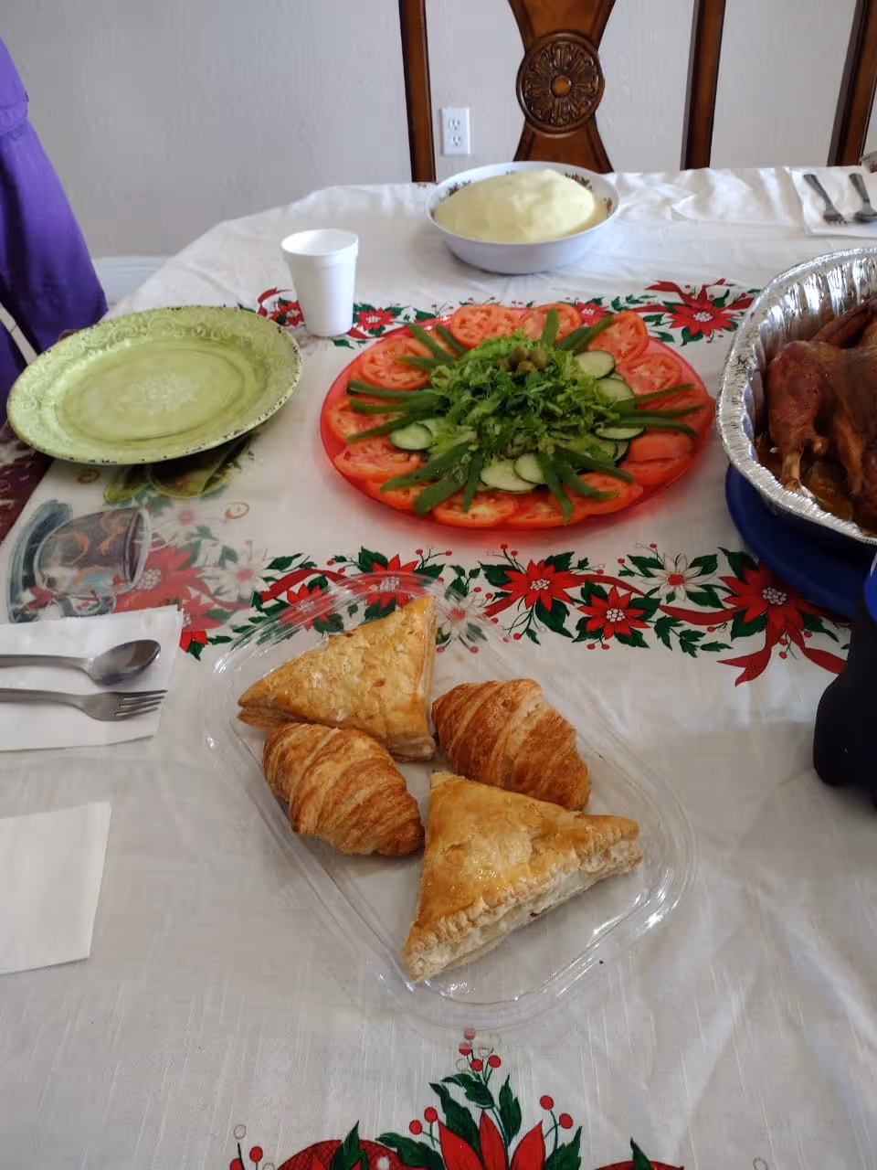 A dining table set with a white tablecloth decorated with red and green poinsettia patterns. On the table, there is a clear plastic tray with two croissants and two triangular pastries, a plate with sliced tomatoes, cucumbers, green beans, and lettuce arranged in a circular pattern, a bowl of mashed potatoes, a green plate, a white disposable cup, and a foil tray with cooked meat. A wooden chair is visible in the background.