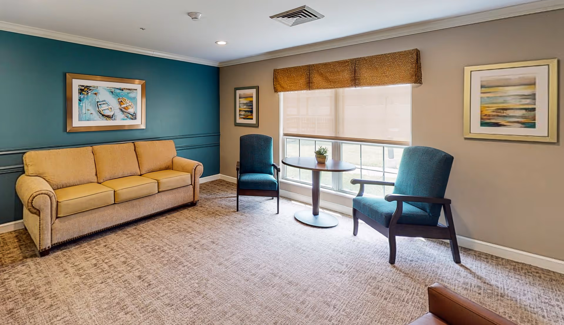 A cozy living room with a beige sofa, two teal armchairs, a round wooden table with a small plant, and three framed paintings on the walls. The room has a large window with a beige roller shade and a brown valance, beige carpet, and teal accent wall.