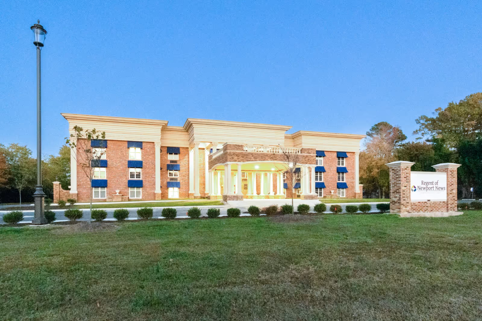 Front exterior of a three-story brick senior living building with columns, a lit entrance, and a lawn sign reading 'Regent of Newport News'.
