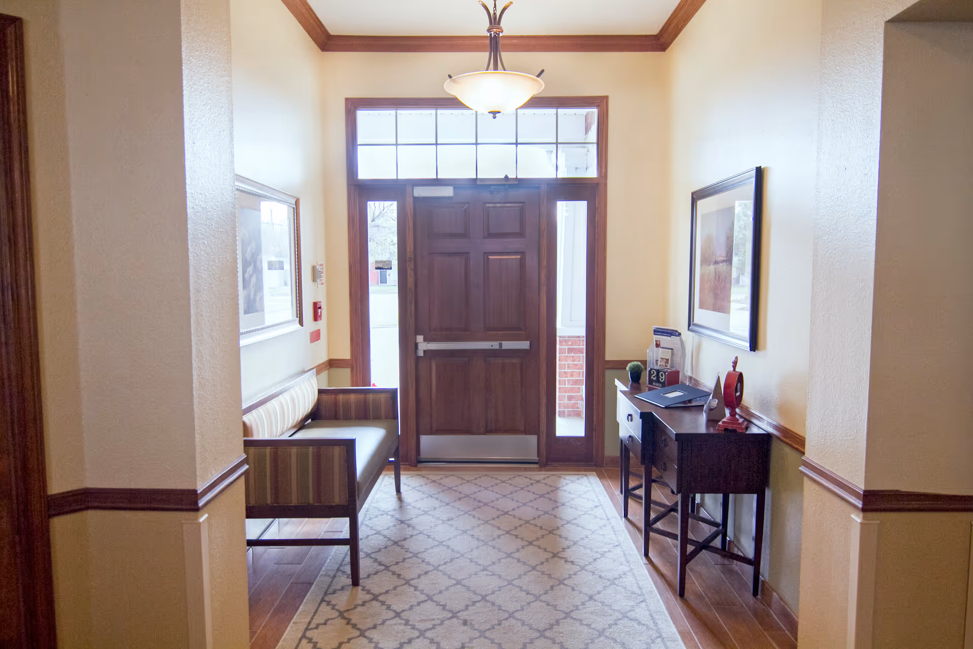 Entrance area of Homestead Assisted Living of Bethany featuring a wooden front door with glass panels above and on the sides, a striped cushioned bench on the left, a wooden console table with decorative items and framed artwork on the right, and a patterned rug on the wooden floor.