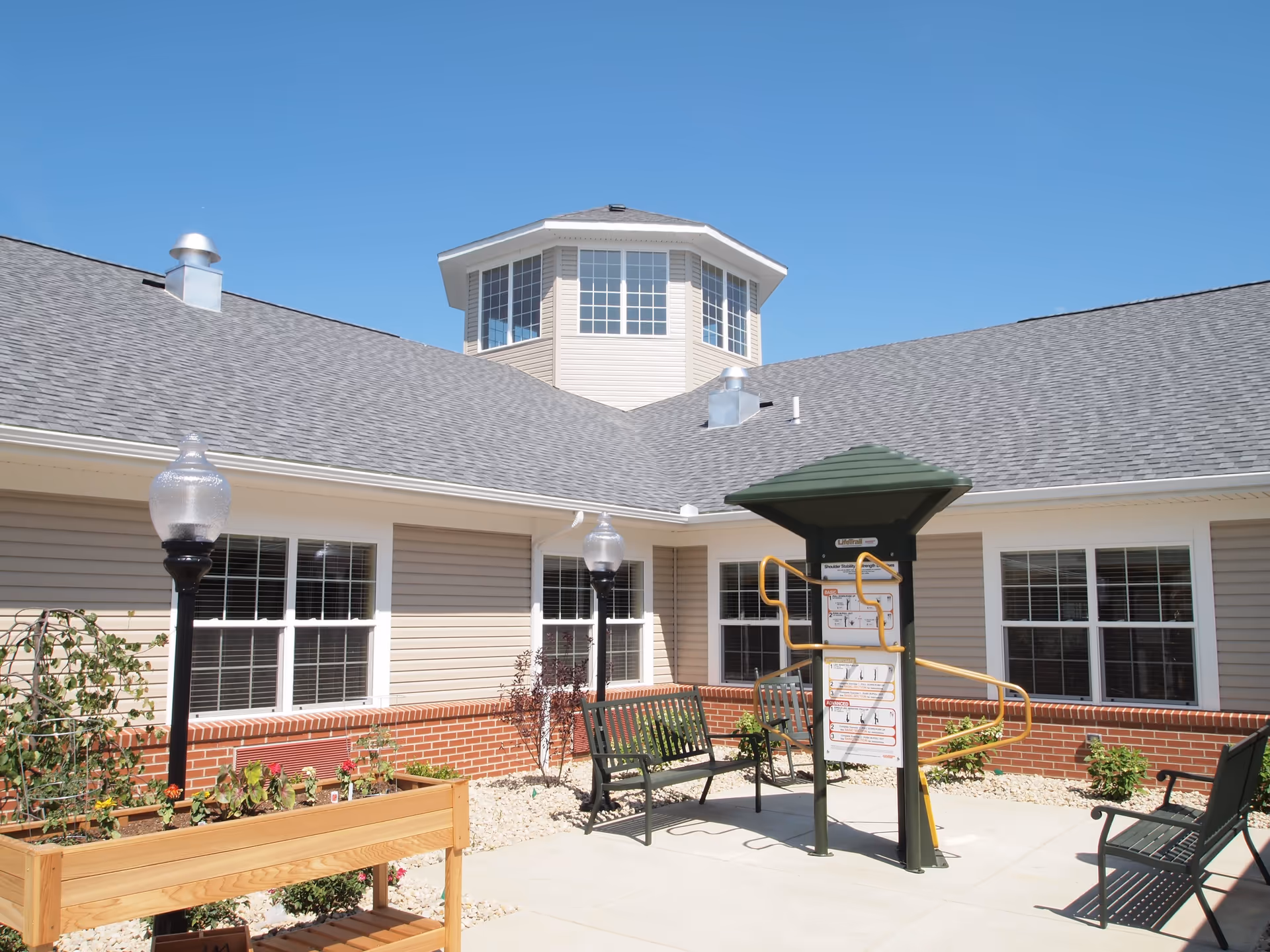 Outdoor courtyard area at The Willows at Willard featuring a paved patio with benches, a raised wooden planter with flowers, and an exercise station under a green canopy. The building surrounding the courtyard has beige siding, red brick accents, and multiple windows under a clear blue sky.