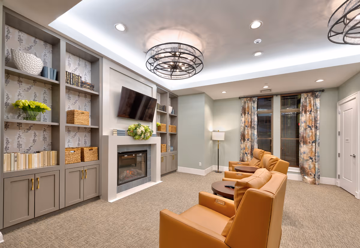 A cozy living room area with three tan leather armchairs facing a wall-mounted flat-screen TV above a modern fireplace. Built-in shelves on either side of the fireplace hold books, baskets, and decorative items. The room has soft lighting from recessed ceiling lights and a floor lamp, with patterned curtains covering the windows.
