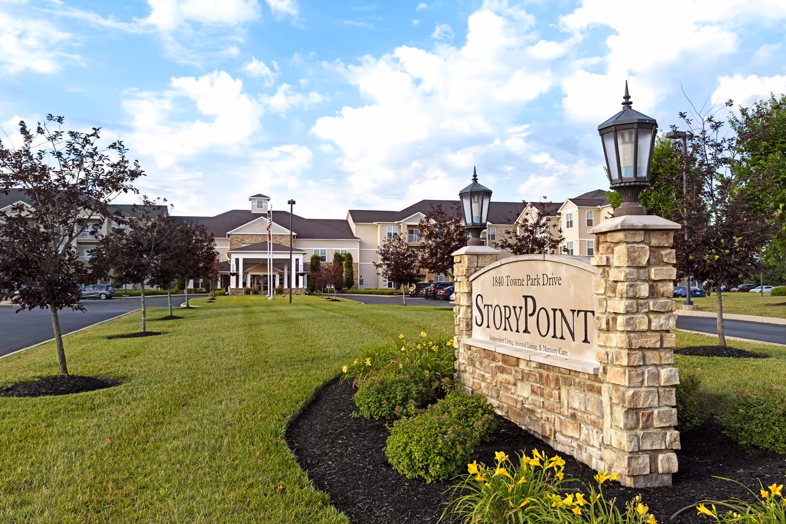 Exterior view of StoryPoint Troy senior living facility showing a large stone sign with the facility name and address, a well-maintained lawn with small trees and flowers, and the building in the background under a partly cloudy sky.