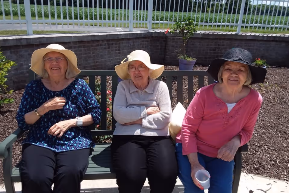 Three elderly women sitting on a green bench outdoors, each wearing a sun hat. They are smiling and appear to be enjoying a sunny day in a garden area with plants and a brick wall behind them.