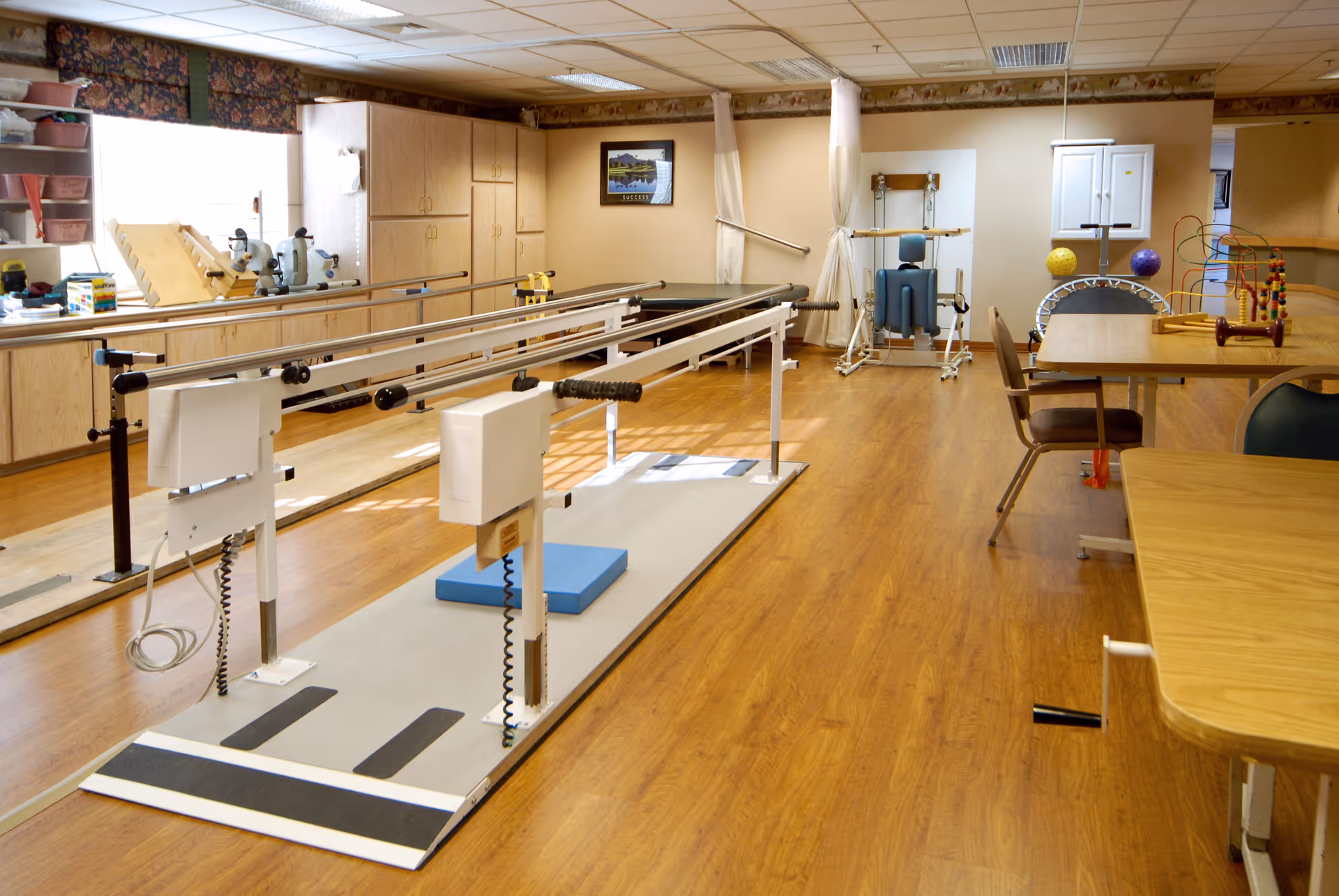 A rehabilitation or physical therapy room with parallel bars for walking exercises, therapy tables, chairs, and various therapy equipment including a small trampoline and colorful activity toys. The room has wooden flooring, beige walls, and cabinets along the back wall.