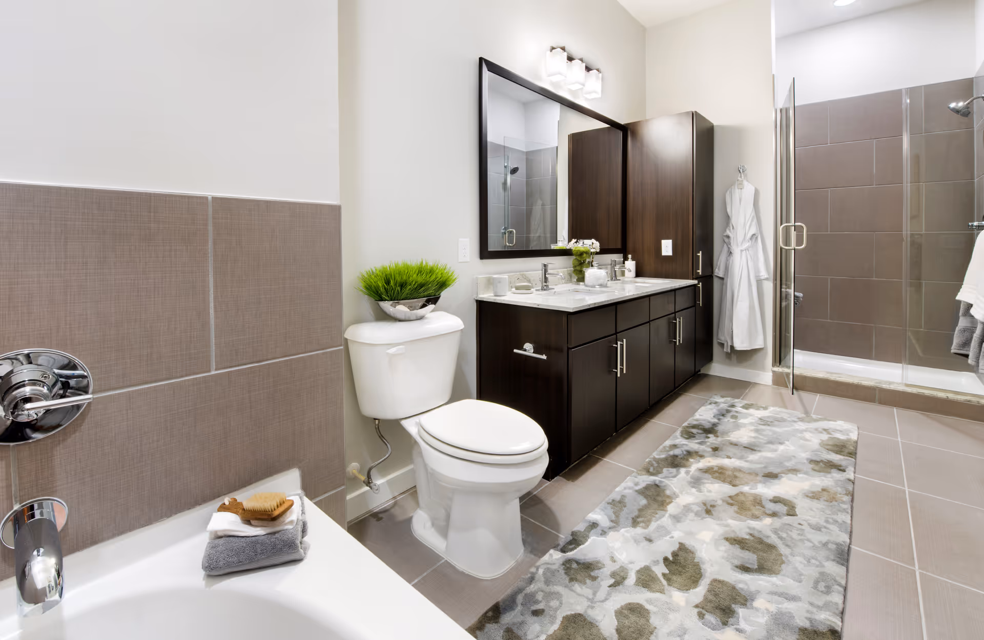 Modern bathroom with a white toilet, dark wood vanity with a large mirror, a glass-enclosed shower, a bathtub with folded towels and a brush, a decorative green plant on the toilet tank, and a patterned rug on the tiled floor.