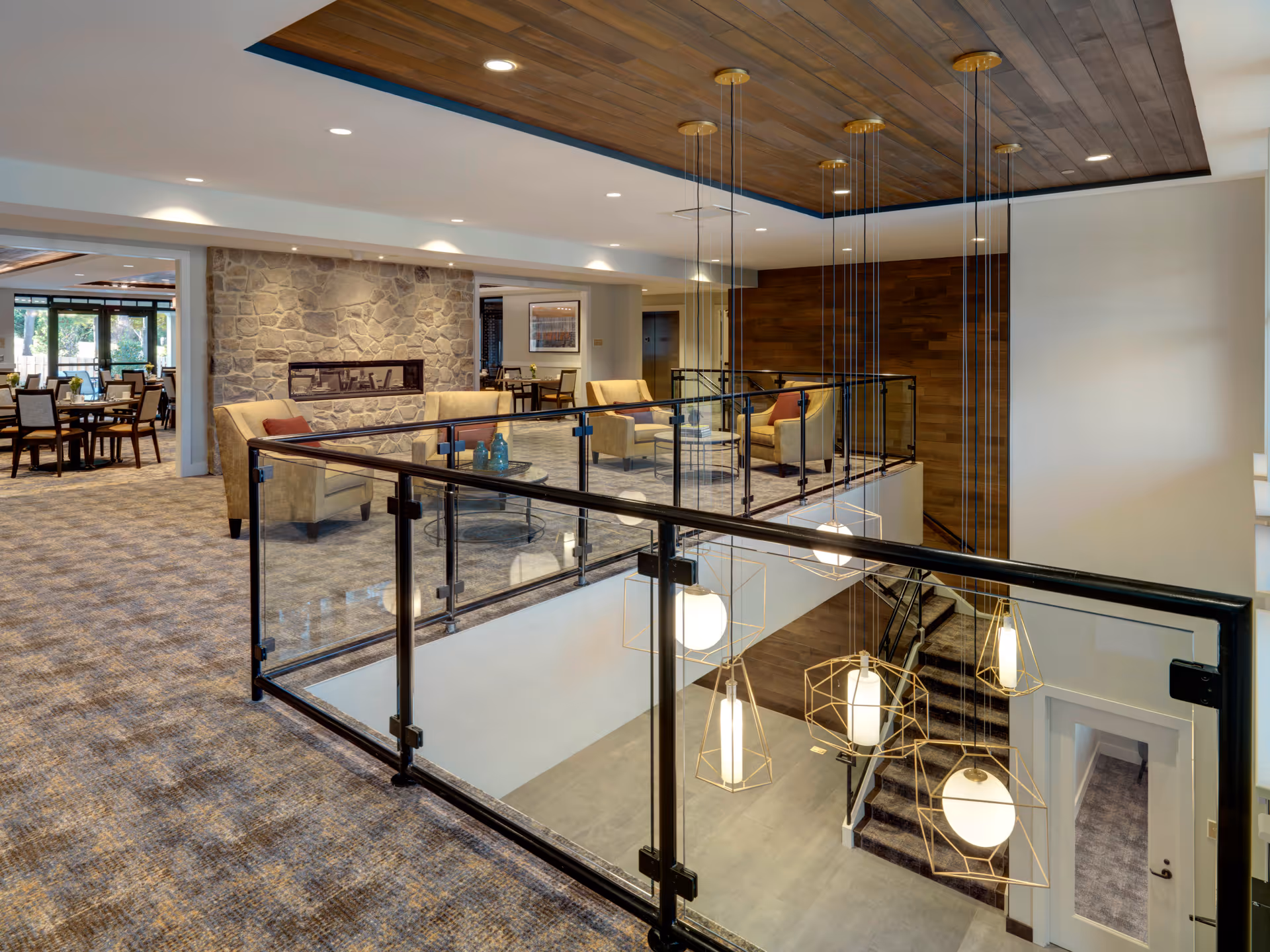 Interior view of a senior living facility featuring a seating area with beige armchairs and a glass coffee table near a stone fireplace. The area overlooks a staircase with modern pendant lights hanging from a wooden ceiling. In the background, there is a dining area with tables and chairs near large windows.