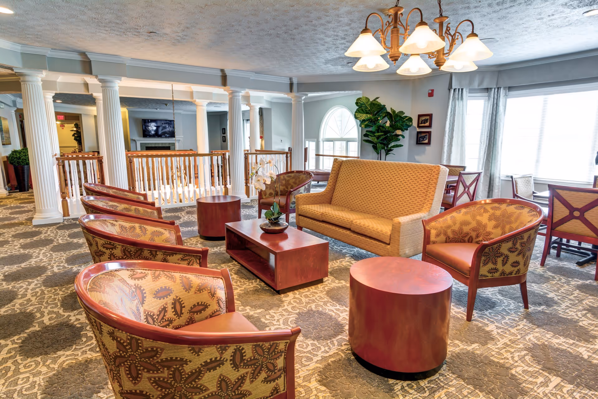 A spacious and well-lit living room area with patterned carpet, a yellow sofa, several patterned armchairs, wooden coffee tables, and a chandelier hanging from the ceiling. Large windows with curtains allow natural light to fill the room. White columns and railings separate this area from another part of the facility.