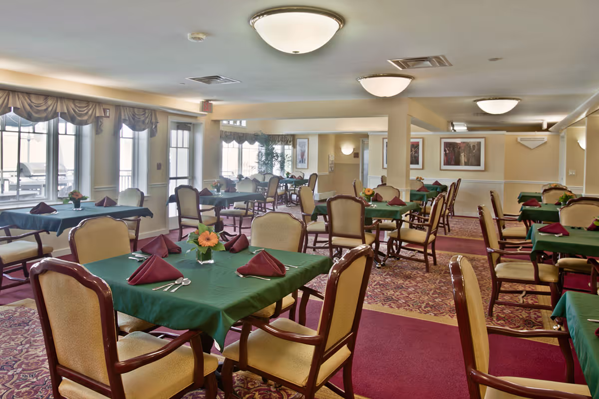 A dining room with multiple tables covered in green tablecloths and set with burgundy napkins, silverware, and small floral centerpieces. The room has beige walls, large windows with valances, and patterned carpet with a red and gold design. Comfortable upholstered chairs with wooden frames surround the tables.